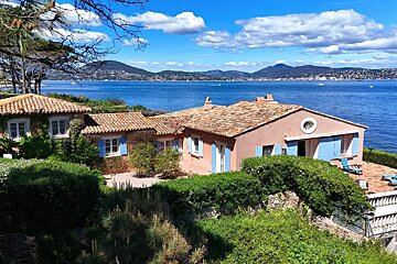 A scenic coastal villa with pink walls and blue shutters sits on a lush hillside overlooking a deep blue bay and distant mountains.
