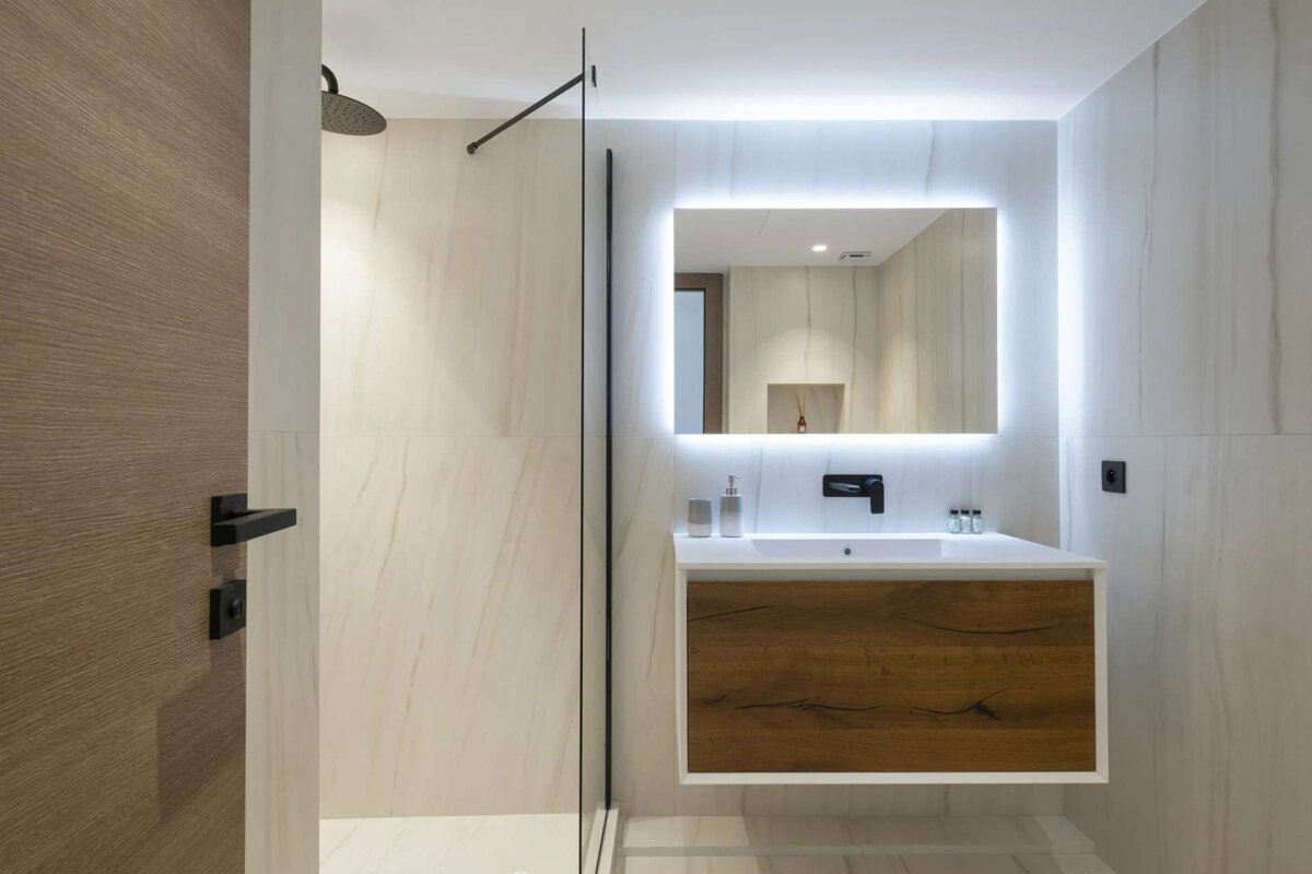 Modern bathroom with a glass shower, floating wooden vanity, and an illuminated mirror. Neutral tones & black fixtures.