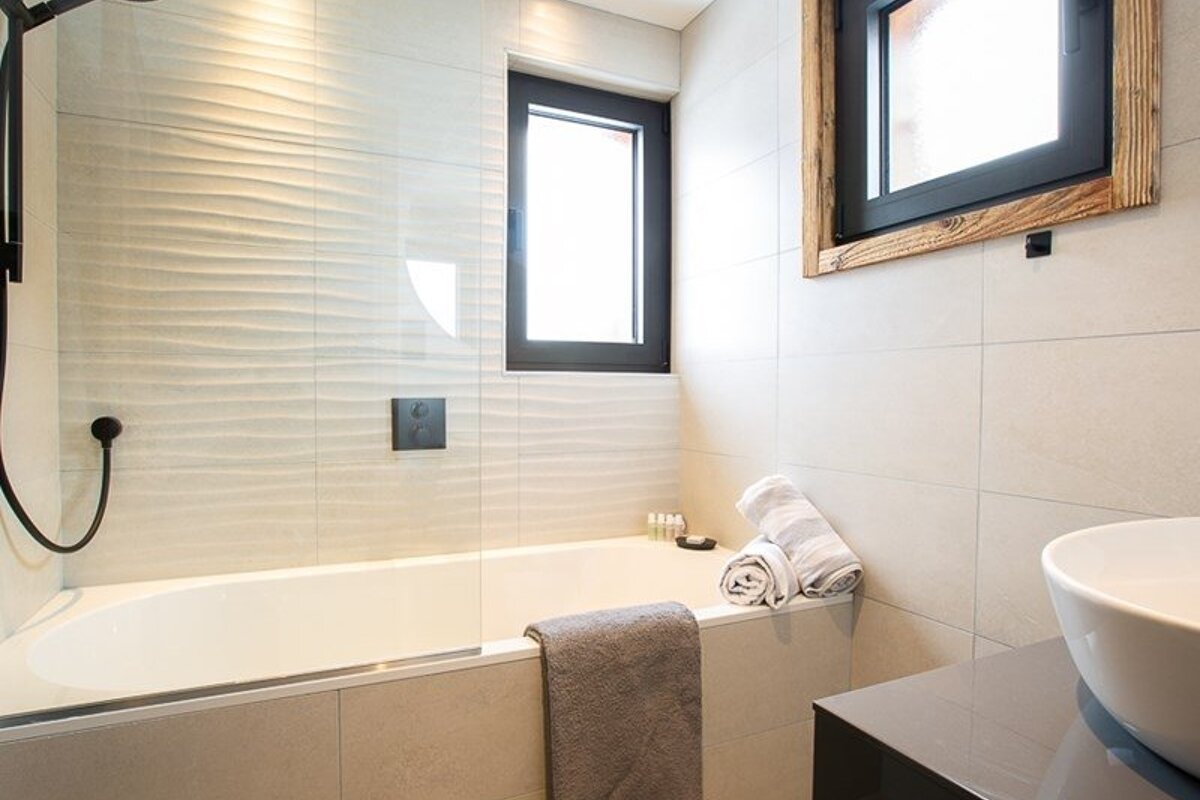 Modern bathroom featuring a white bathtub, glass partition, textured beige tiling, and black-framed windows.