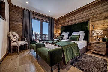 A luxurious, wood-paneled chalet bedroom with a plush green velvet bed, deer-themed decor, and a scenic balcony overlooking snow-capped mountains.