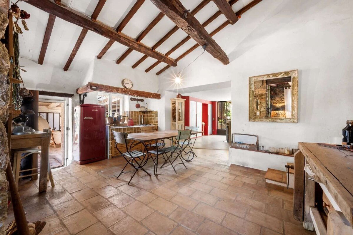 A charming rustic kitchen with exposed wood beams, terracotta tiles, white walls, a vintage red fridge, and a dining table.