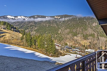 Balcony view of a snowy mountain slope, evergreen forests, and a small village nestled in the valley under a bright blue sky.