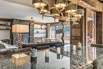 A luxurious, rustic-modern chalet interior blends wood, stone, and fur accents. A unique pebble-under-glass table overlooks snowy mountain views.