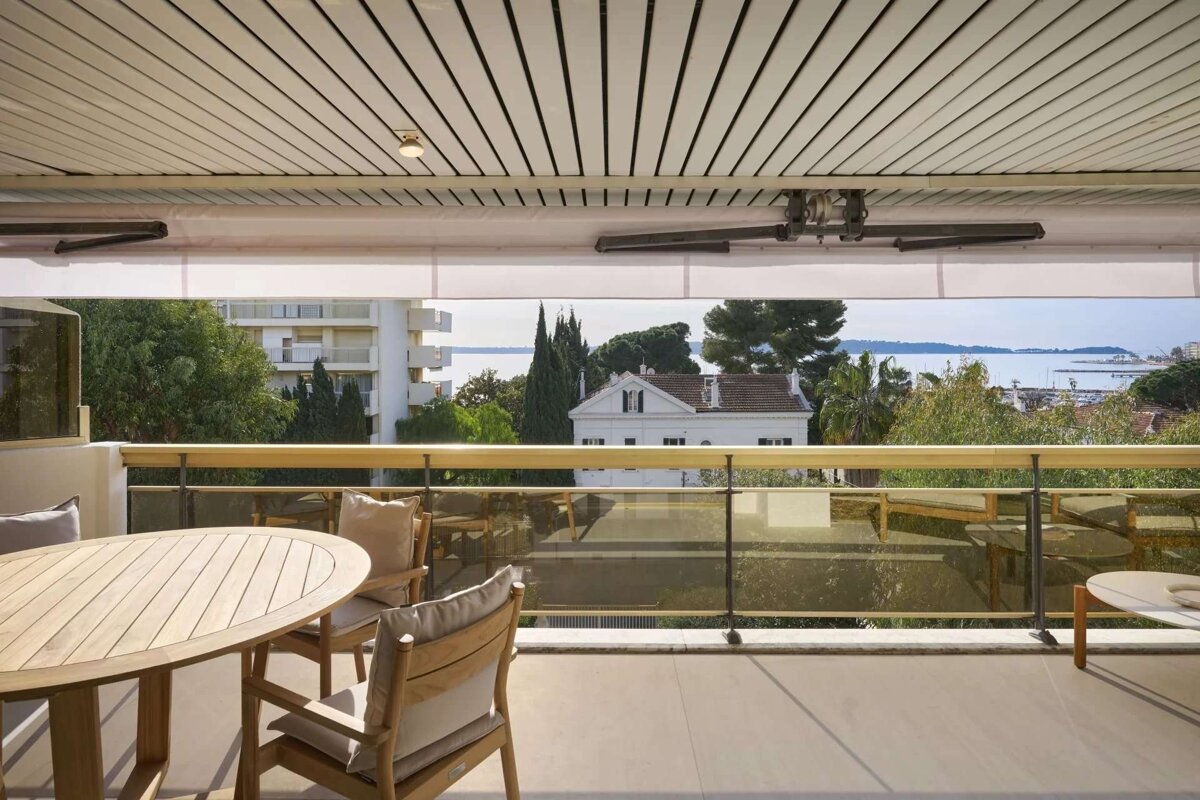 A balcony with wooden patio furniture, providing a scenic view of white buildings, lush trees, and the ocean beyond.