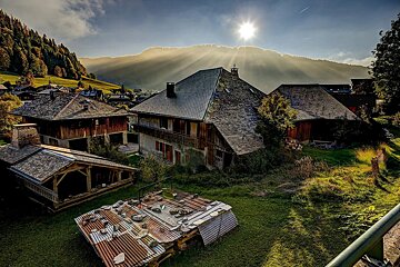 Golden sunburst over a rustic mountain village. Old houses with steep roofs, green hills, and corrugated metal in the foreground bathed in light.