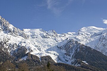 Stunning view of snow-capped mountains with a massive glacier cascading down, bordered by dark forests, under a brilliant blue sky.