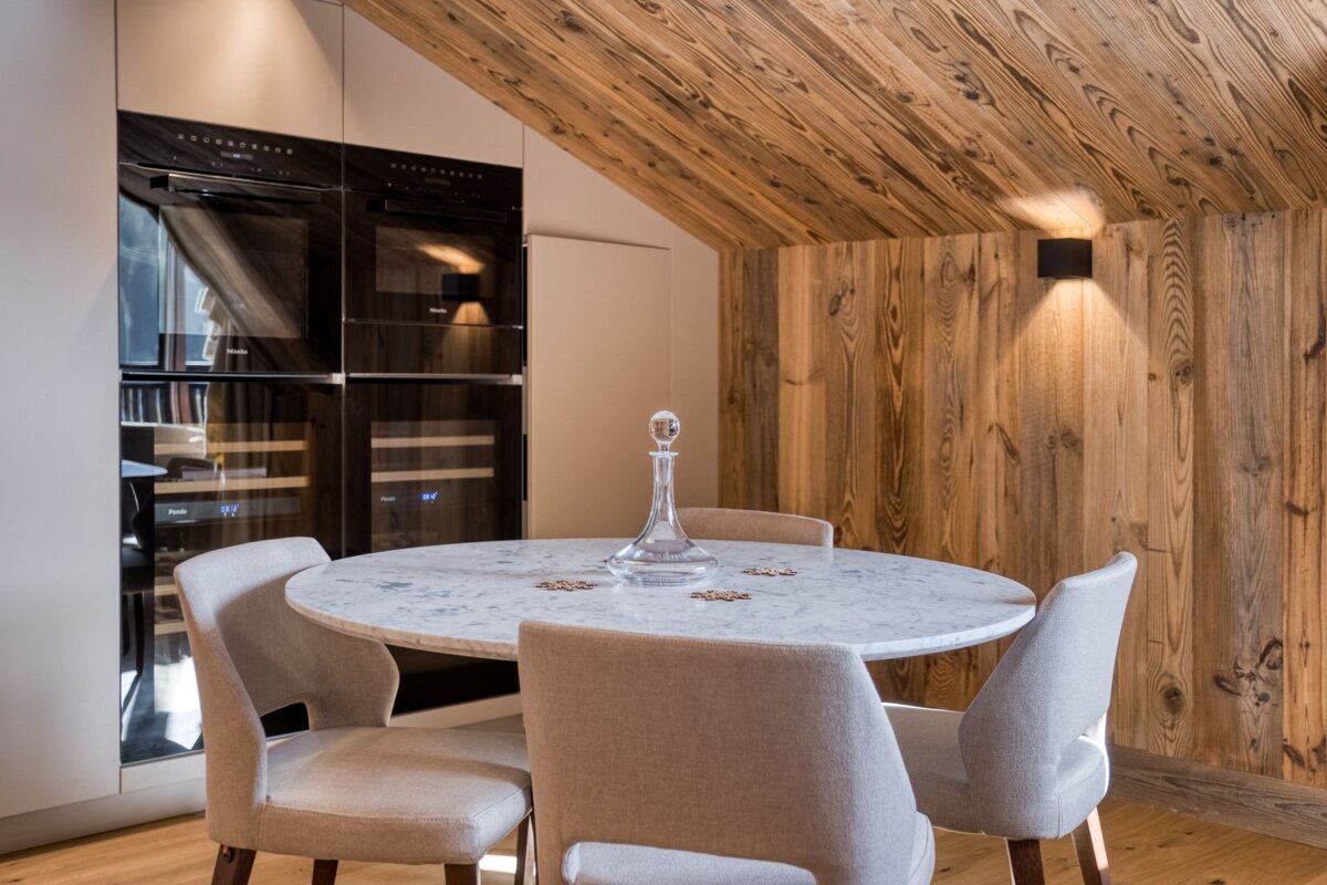 A modern kitchen/dining area features a marble table, gray chairs, built-in appliances, and warm wood paneling on the walls and sloped ceiling.