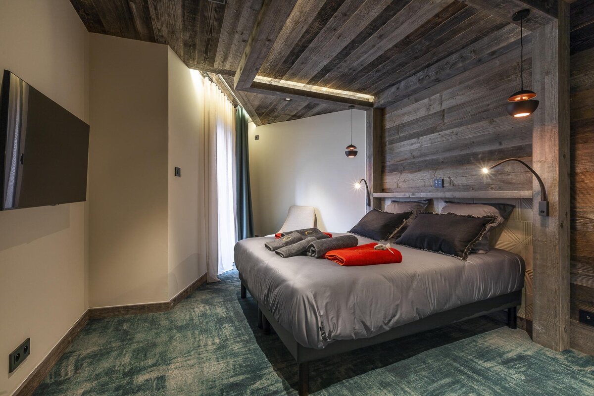 A cozy modern bedroom with rustic wood paneled walls and ceiling, a grey bed, a wall-mounted TV, and patterned green carpet.