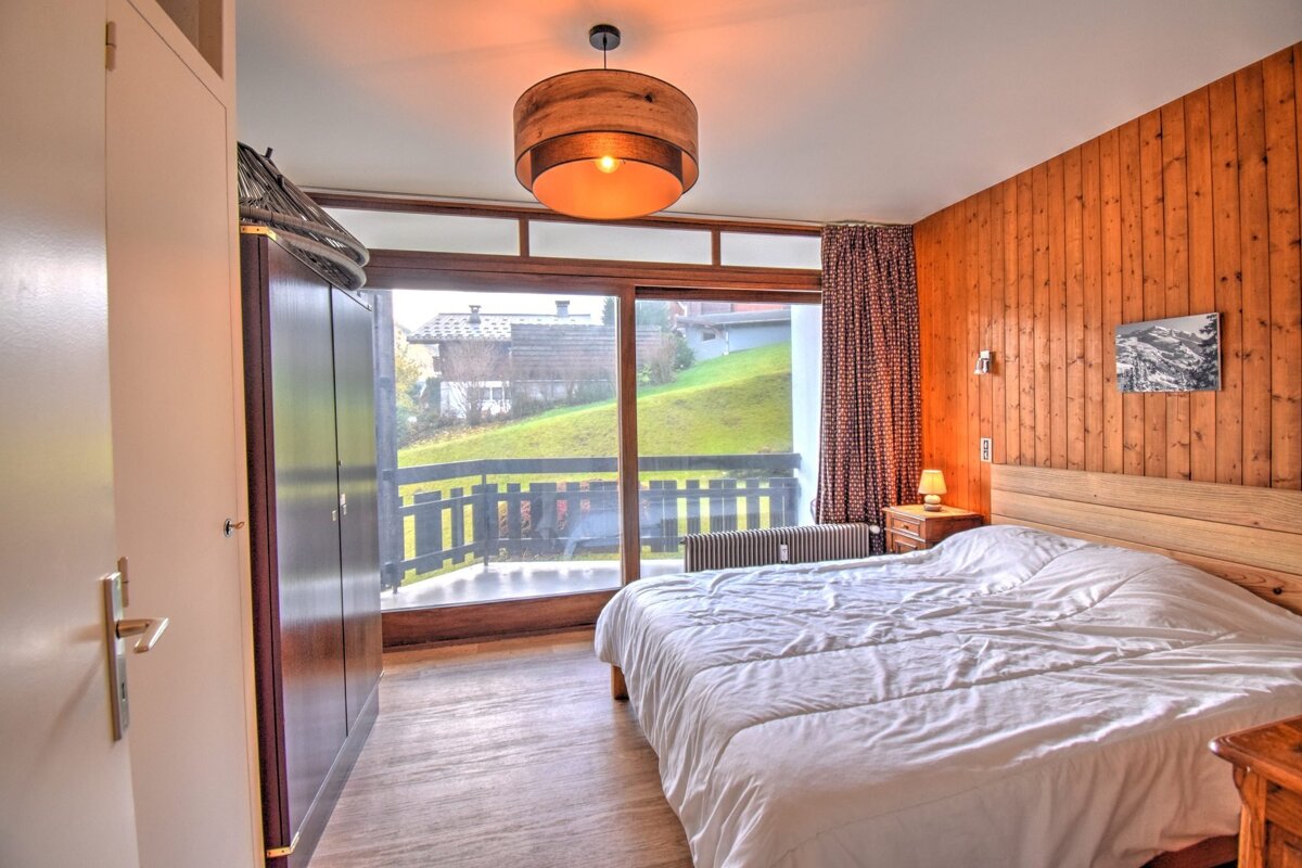 Cozy cabin-style bedroom with a wood-paneled wall, large bed, and sliding glass doors opening to a scenic mountain-view balcony.