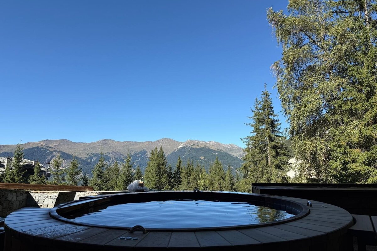 An outdoor wooden hot tub overlooking a pine forest and majestic mountains under a clear blue sky.