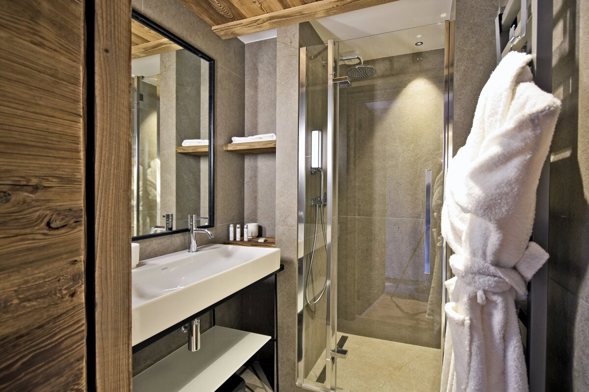 A modern bathroom featuring a white sink, large mirror, glass shower cabin, and a white bathrobe on a towel warmer. It has tiled walls and wood accents.