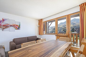 A cozy living/dining room with a wooden table, brown sofa, and abstract art. Large windows frame a stunning mountain village view.