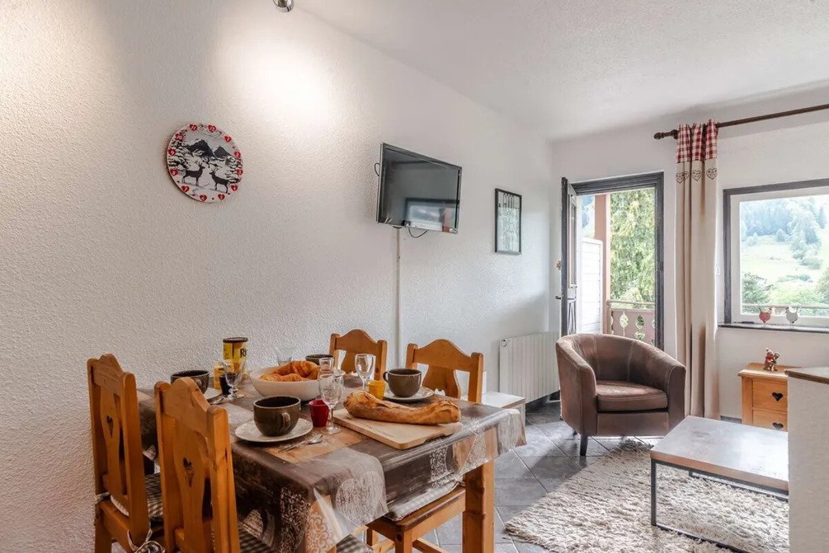 A bright room with a set breakfast table, wooden chairs, TV, and an armchair. A large window and balcony door reveal a scenic green, hilly view.