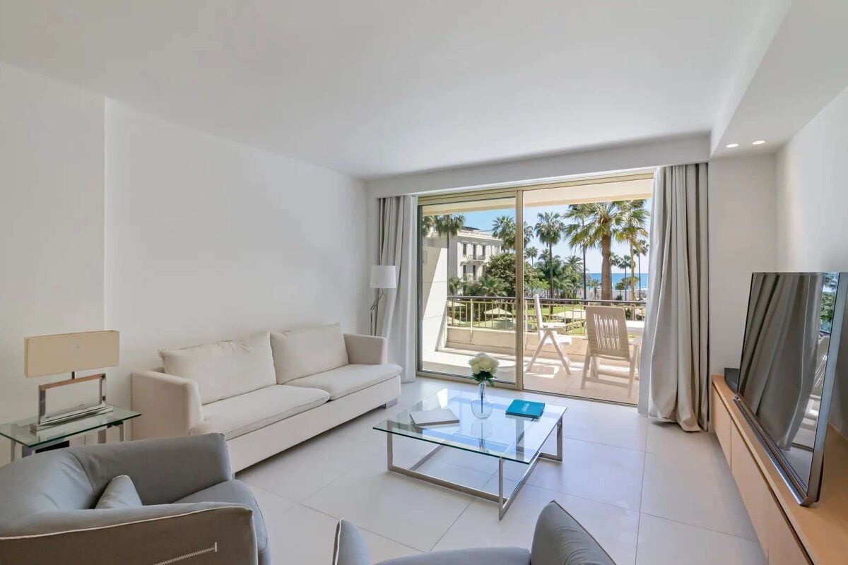 Bright, modern living room with white sofa, glass table, and balcony overlooking palm trees, a building, and the sea.