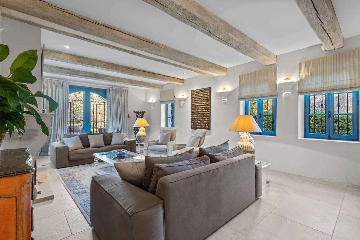 A bright living room with exposed wooden beams, white walls, and grey sofas. Features striking blue-framed windows/door, tiled floors, and warm lamps.