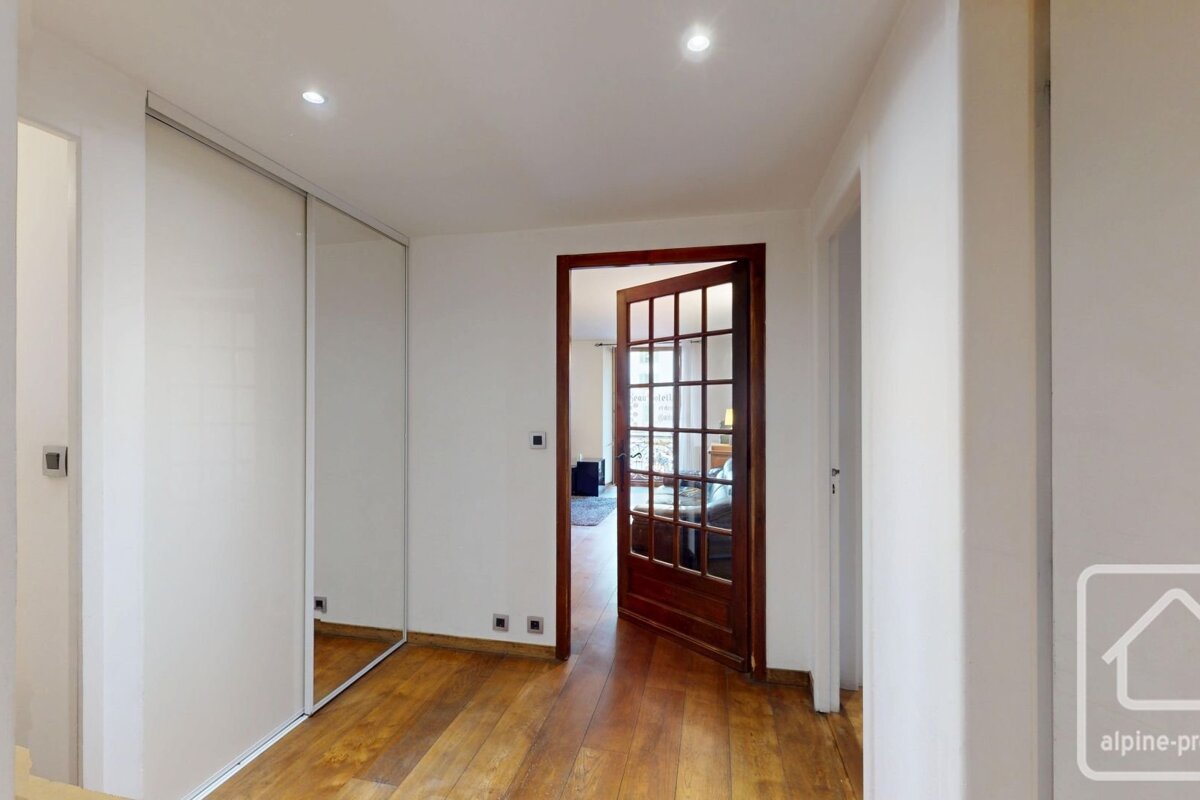A bright hallway with a warm wooden floor, white walls, a sliding closet, and an open dark wood glass-paned door leading to another room.