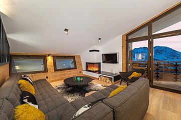 A cozy alpine living room features a lit corner fireplace, large grey sofa with yellow cushions, and expansive mountain views through full-height windows.