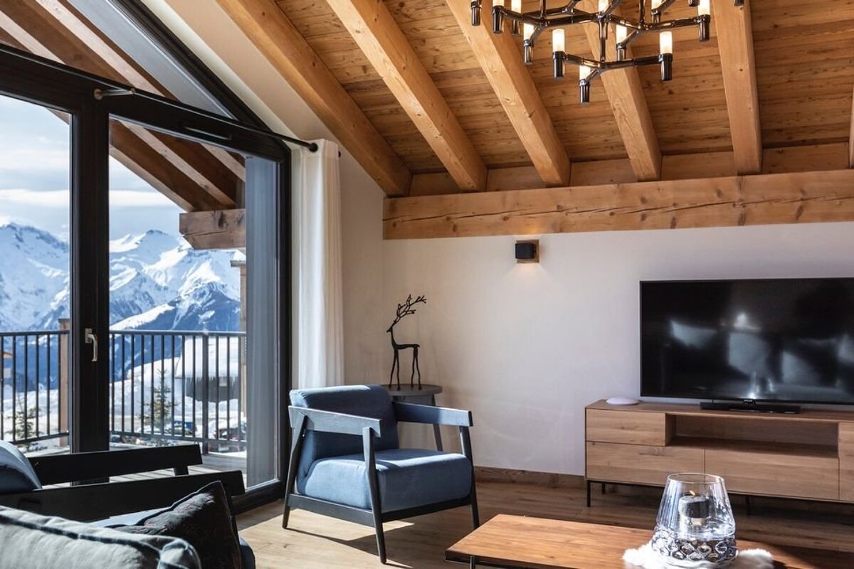 Cozy chalet living room with wooden beams, a blue armchair, and a stunning snowy mountain view through large glass doors.