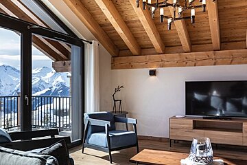 Cozy chalet living room with wooden beams, a blue armchair, and a stunning snowy mountain view through large glass doors.