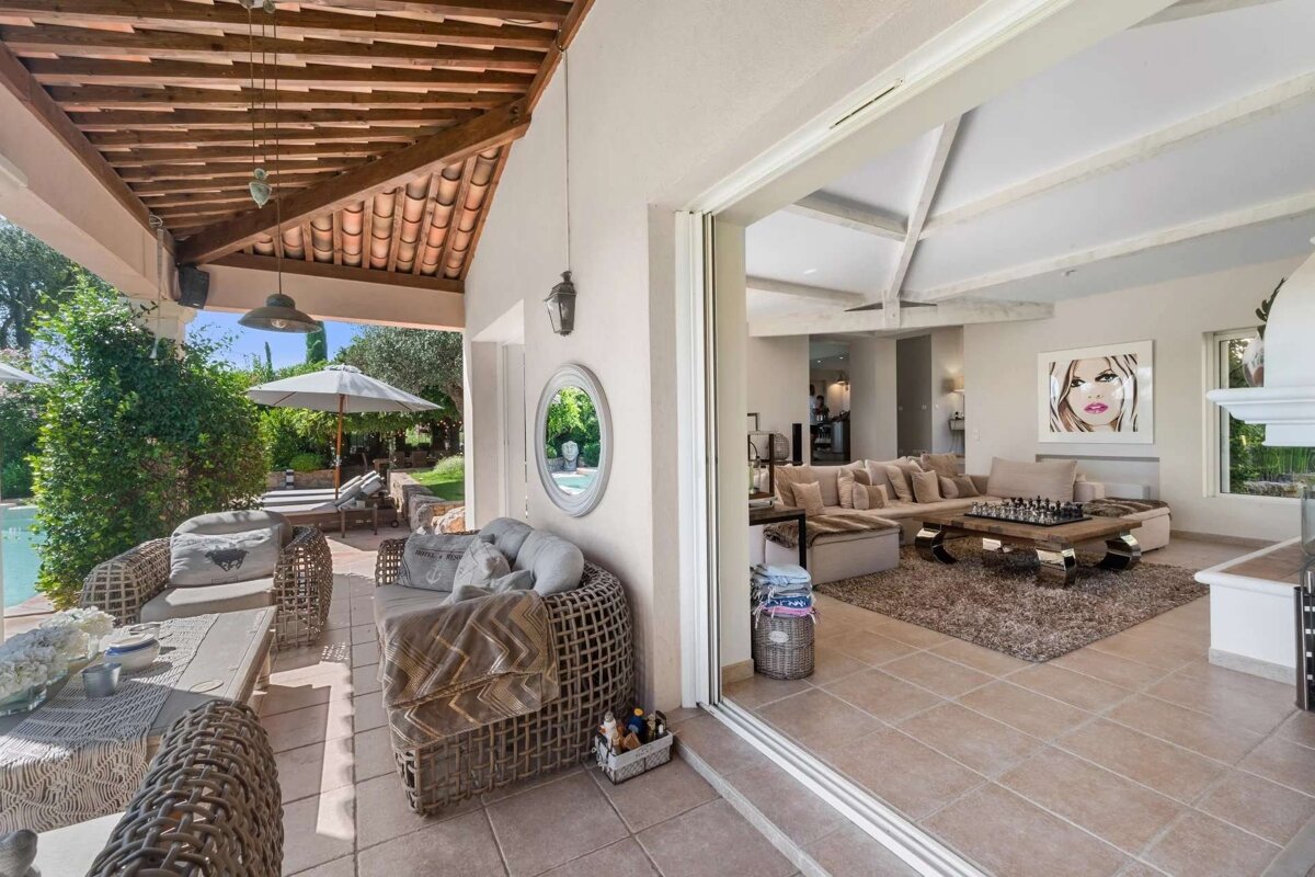 An inviting open-plan home featuring a stylish indoor living room and a covered outdoor patio with wicker seating, overlooking a pool and garden.