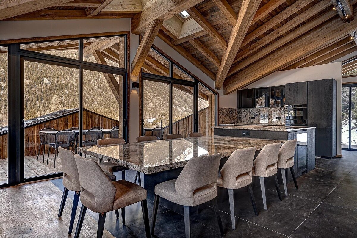 Modern luxury chalet dining room with vaulted wooden ceilings, marble surfaces, and scenic mountain views through large windows.