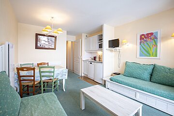A bright, cozy living room features a dining area with mismatched chairs, two teal sofas, a compact white kitchen, and cream walls with art.