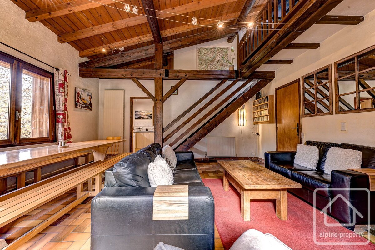 A rustic living room with dark leather sofas, wooden beams, a staircase, and a dining table.