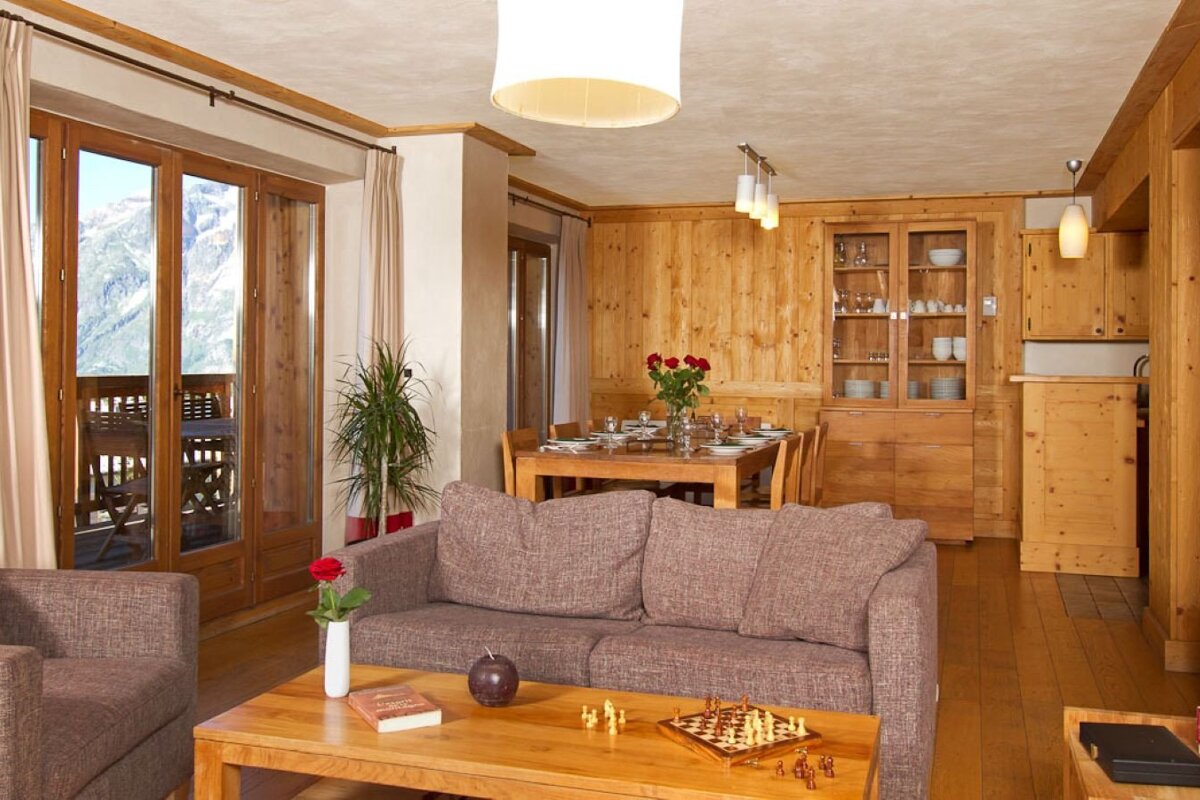 Cozy, wood-paneled living/dining room with a mountain view from the balcony. Features a sofa, coffee table with chess, and a set dining table.