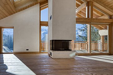 Modern chalet interior with a central fireplace, wooden accents, and large windows revealing a stunning snowy mountain landscape.