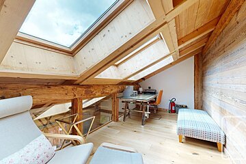 A warm, bright attic space with exposed wooden beams, walls, and floor. Skylights illuminate a work desk, office chair, and lounge seating.