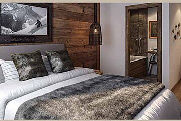 Cozy rustic bedroom with wood paneled walls, a furry grey blanket, and an open doorway leading to a tiled bathroom with a tub.
