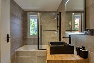 A modern bathroom with neutral grey tiles, a tub/shower combo, window to trees, and a black stone vessel sink on a light wood vanity.