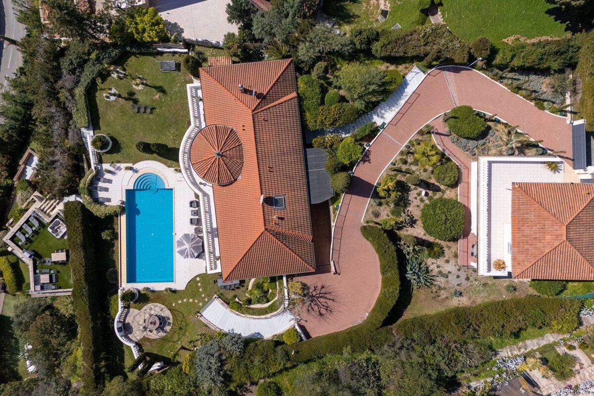 Aerial view of a luxury villa with a terracotta roof, a bright blue swimming pool, a winding driveway, and lush green gardens.