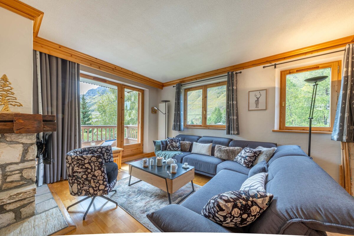 Cozy living room with a large blue sectional sofa, stone fireplace, and mountain views from large windows and a balcony. Wood accents create a warm feel.