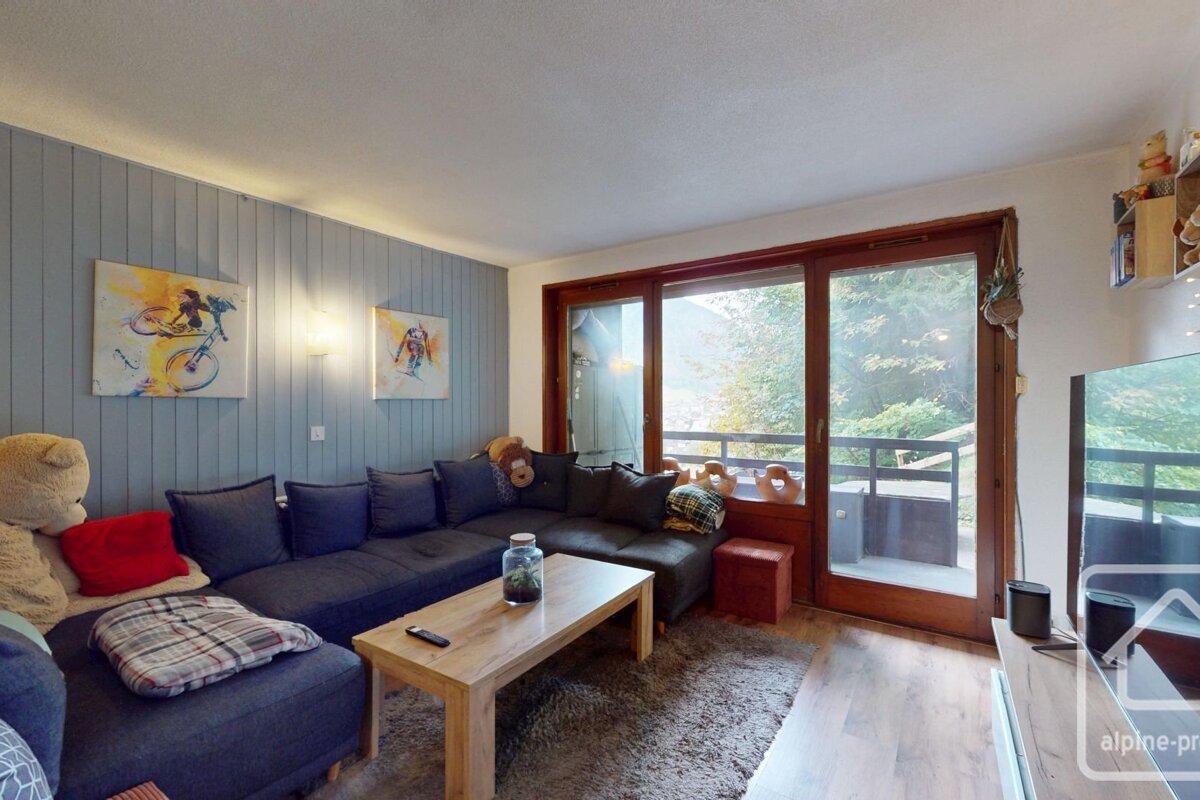 A cozy living room features a large sectional sofa, wooden coffee table, colorful art on a paneled wall, and a balcony overlooking nature.