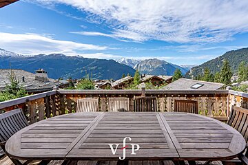 Wooden dining table and chairs on a balcony with a panoramic view of mountain peaks and village chalets.