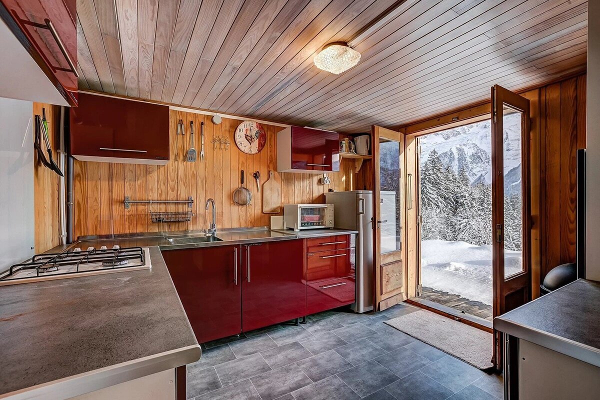 A rustic kitchen with wood paneling and red cabinets opens to a snowy mountain landscape with pine trees.