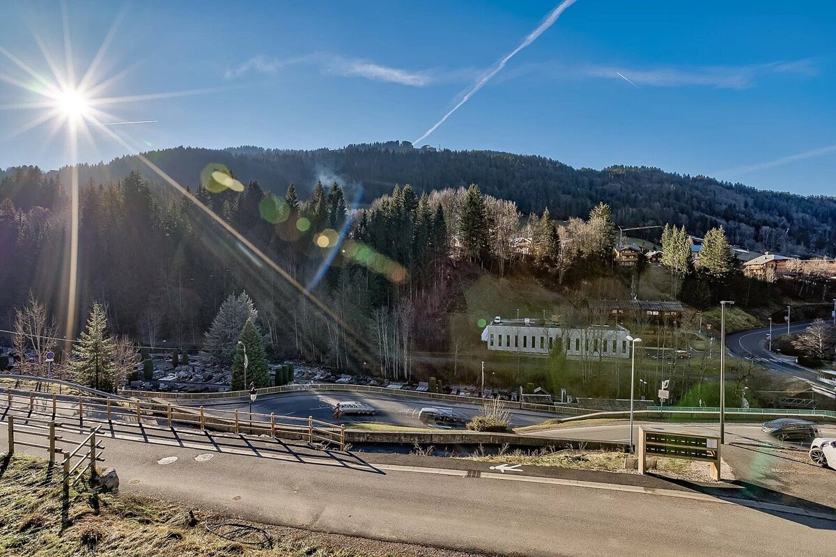 Sunny mountain landscape with dense forests, scattered buildings, and a winding road under a bright blue sky with lens flare.