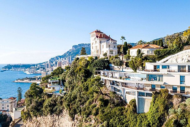 Luxurious villas and modern homes cascade down a lush green hillside, overlooking a bustling coastal city and sparkling blue sea under a clear sky.