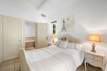 A bright, white bedroom features a double bed with a rattan headboard, two nightstands with lamps, and Eiffel Tower art. An open door reveals a bathroom.
