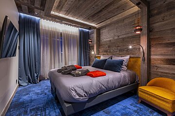 Chic bedroom with rustic wood paneling, vibrant blue carpet, a large bed with modern linens, and warm accent lighting. A yellow armchair adds a pop of color.