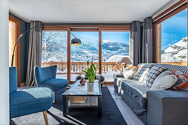A cozy living room with large windows framing a vast snowy mountain vista. Features blue armchairs, a grey sofa, coffee table, and an arc lamp.