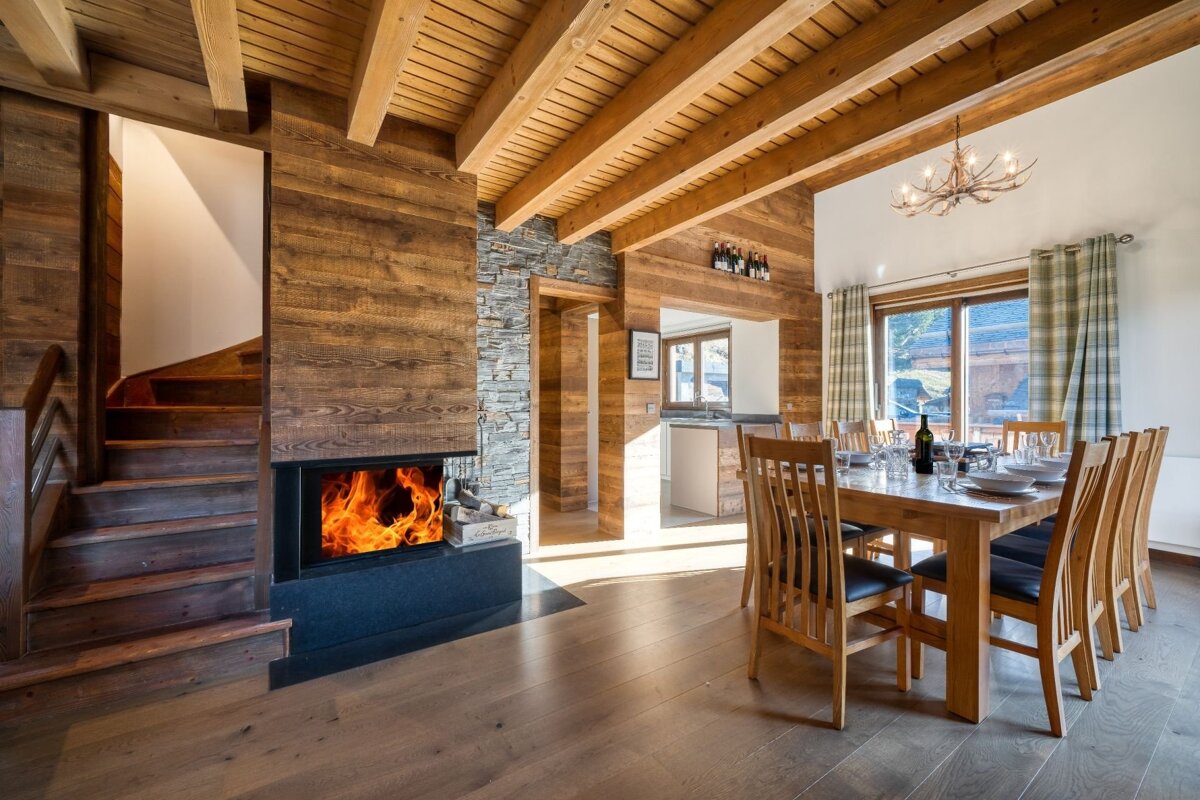 Warm chalet interior featuring a modern fireplace, wooden beams, dining area, and stairs. Natural light fills the cozy, rustic-modern space.