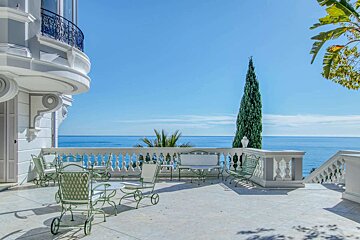 Ornate seaside terrace with green patio furniture, offering stunning blue ocean views under a clear sky, featuring a tall cypress tree.
