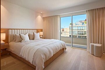 Serene, modern bedroom in neutral tones with a large bed, light wood floors, and a balcony view through large glass doors.