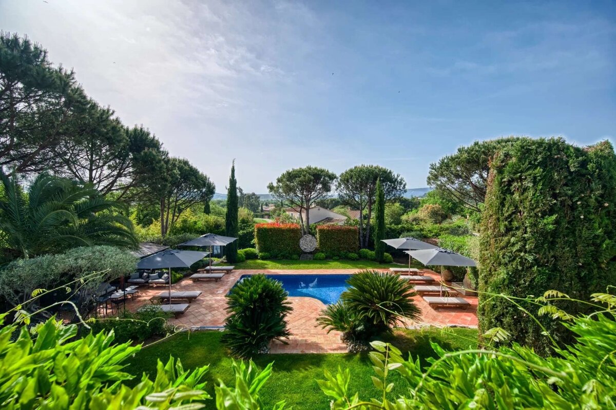 A beautiful outdoor swimming pool area surrounded by lush green foliage, lounge chairs, and umbrellas under a sunny blue sky.