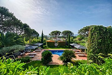 A beautiful outdoor swimming pool area surrounded by lush green foliage, lounge chairs, and umbrellas under a sunny blue sky.