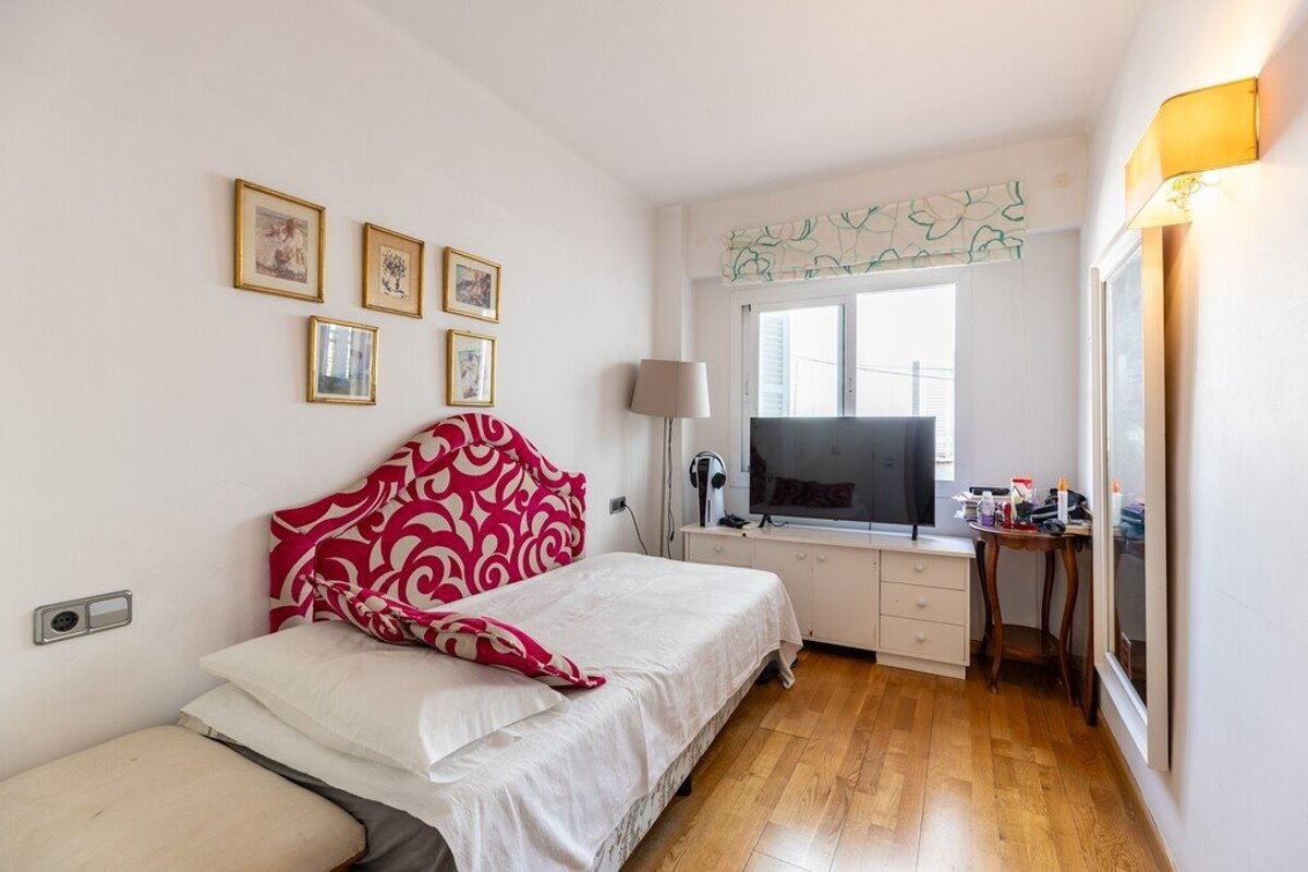 A small bedroom featuring a single bed with a bold pink headboard, hardwood floors, wall-mounted art, and a TV console by the window.