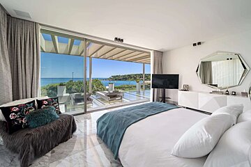 A luxurious modern bedroom with a large white bed, a fur accent chair, and expansive glass doors offering a stunning panoramic sea view.