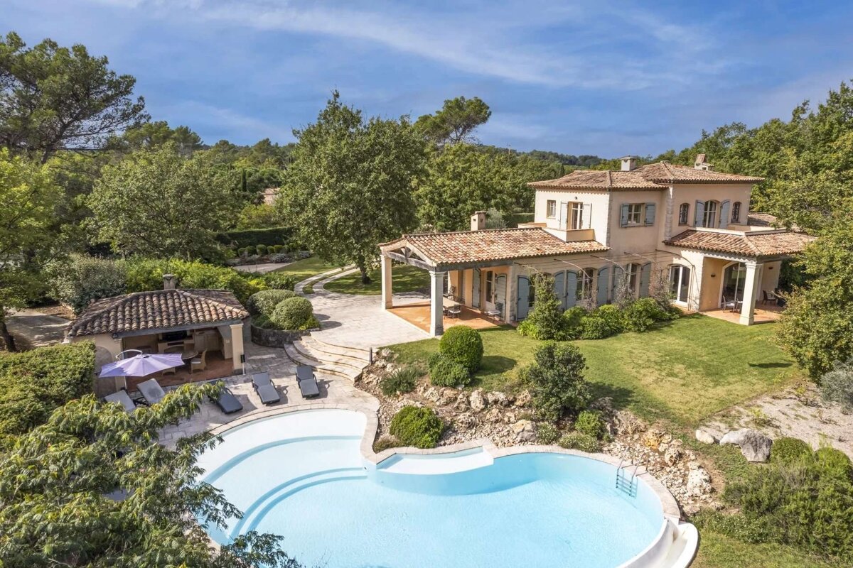 An aerial view of a luxurious Mediterranean-style villa featuring a large, irregular swimming pool, a pool house, and lush green gardens under a clear blue sky.